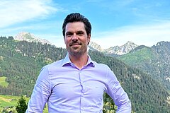 Porträt einer Person in hellem Hemd vor einer alpinen Landschaft mit bewaldeten Hängen, grünen Wiesen und markanten Berggipfeln unter blauem Himmel.