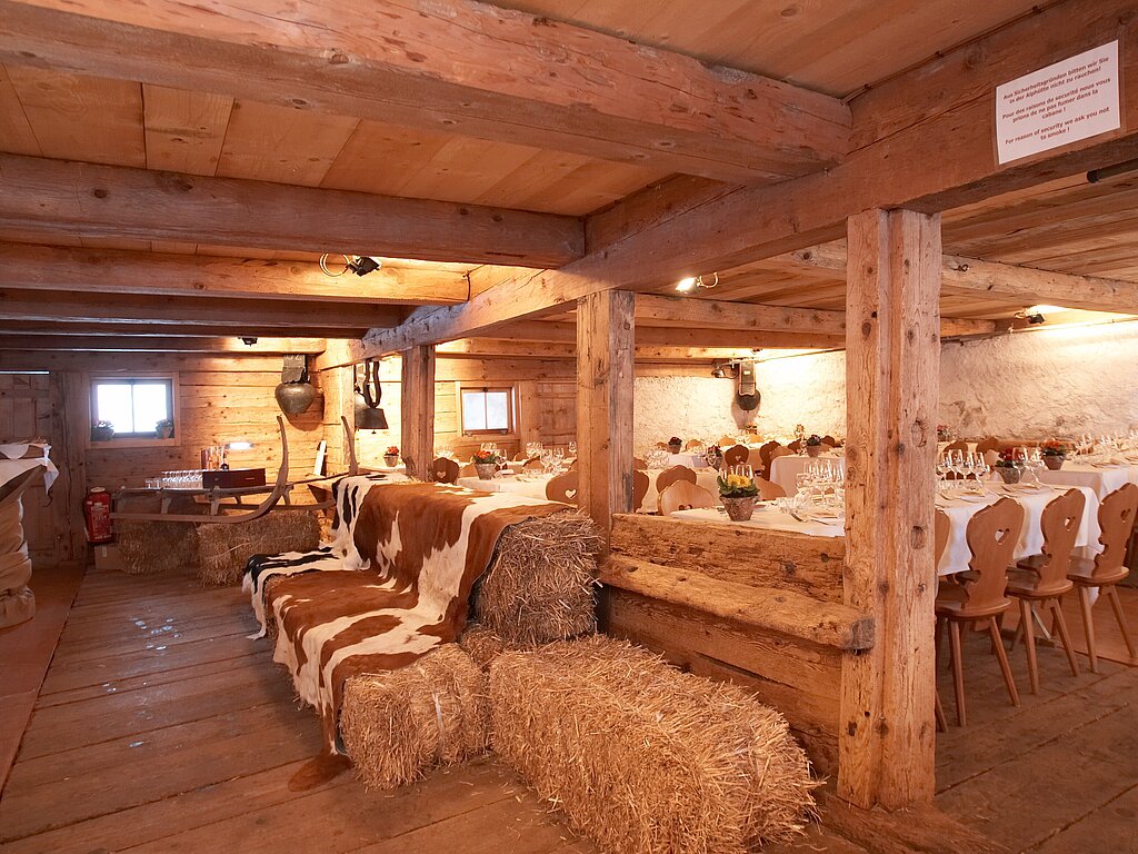 Mit weissen Tischtüchern gedeckte Tische, Holzstühle sowie Strohballen und einem Hornschlitten mit Gläsern in einem rustikalen Stall in einer Alphütte.