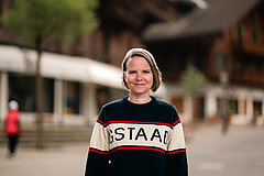 Personne debout à l’extérieur dans un village, portant un pull avec l’inscription « GSTAAD » ; bâtiments traditionnels visibles en arrière‑plan flou.