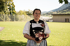 Personne en tenue traditionnelle tenant un bouquet de fleurs sur une prairie verte, avec des bâtiments et des arbres en arrière‑plan.