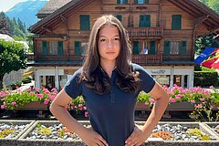 Porträt einer Person vor einem traditionellen Holzchalet. Im Vordergrund befinden sich Blumenbeete mit pinken Blüten, im Hintergrund sind Berge und weitere alpine Elemente zu sehen.