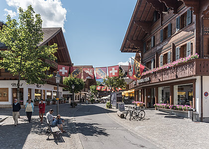 Promenade mit blumengeschmückten Chalets, Fussgängern, einem Mann und einem Kind auf einer Sitzbank, Bäume und Strassenlaternen, in der Mitte hängen Fahnen von der Schweiz, dem Kanton Bern und der Gemeinde Saanen.