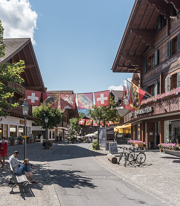 Promenade mit blumengeschmückten Chalets, Fussgängern, einem Mann und einem Kind auf einer Sitzbank, Bäume und Strassenlaternen, in der Mitte hängen Fahnen von der Schweiz, dem Kanton Bern und der Gemeinde Saanen.