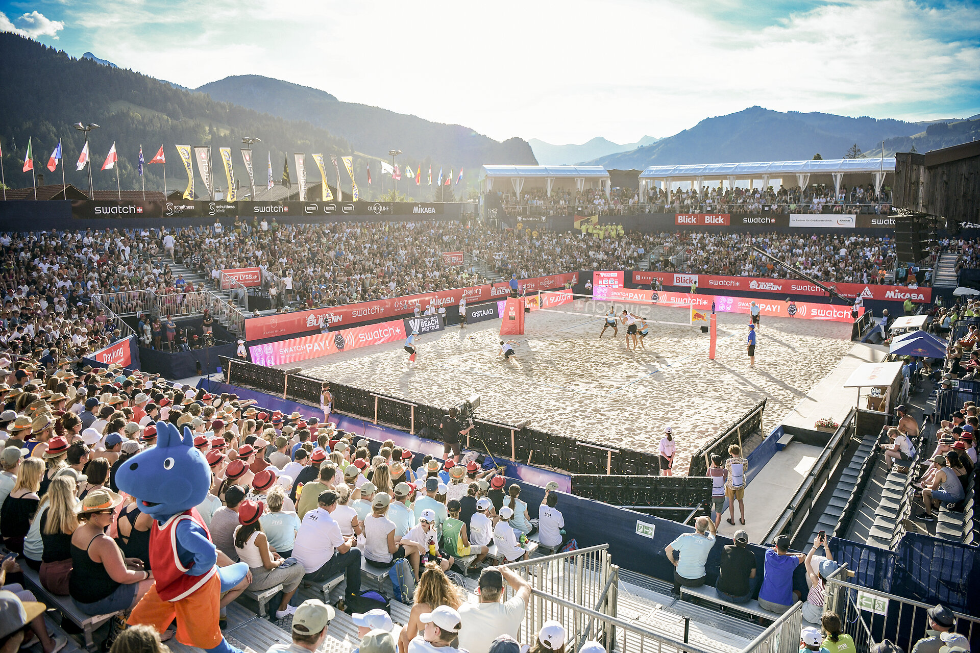 Sicht aufs Spielfeld des Swatch Beach Pro Gstaad, drumherum viel Publikum in den Sitzen. Vorne links ist das Maskottchen zu sehen, im Hintergrund Berge.