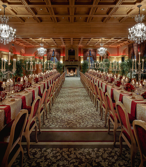 In einem luxuriösen Saal steht ein langer Tisch mit Kerzen, Weingläsern und Blumen.