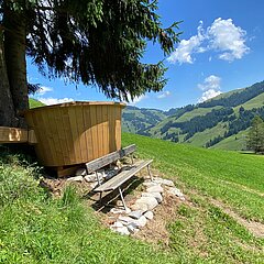 Ein überdimensionales Fondue-Caquelon steht auf einer Wiese in Abländschen. Im Hintergrund sind Berge, ein Bauernhaus und eine sonnige alpine Landschaft zu sehen.