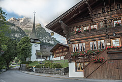 Im Vordergrund sieht man das Hotel Bären in Gsteig. Dies ist ein sehr altes Holzchalet mit roten Geranien vor den Fenstern und alten Zeichnungen an der Front. Im Hintergrund ist die Kirche Gsteig sichtbar.