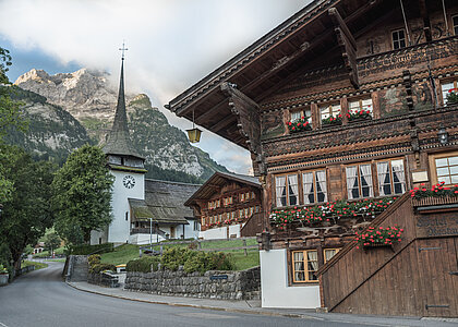 Im Vordergrund sieht man das Hotel Bären in Gsteig. Dies ist ein sehr altes Holzchalet mit roten Geranien vor den Fenstern und alten Zeichnungen an der Front. Im Hintergrund ist die Kirche Gsteig sichtbar.